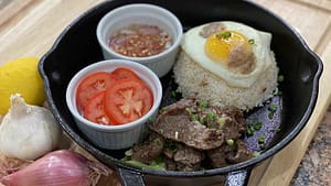 Beef Tapa prepared in an iron skillet with a side of rice with an egg over the top