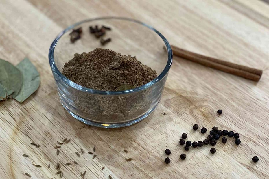 Garam Masala in a cup with ingredients used to make it on wood cutting board