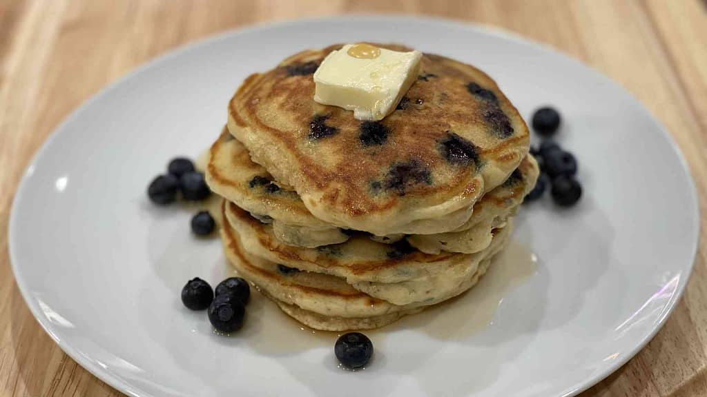 A stack of blueberry pancakes on a plate with butter and raw blueberries surrounding