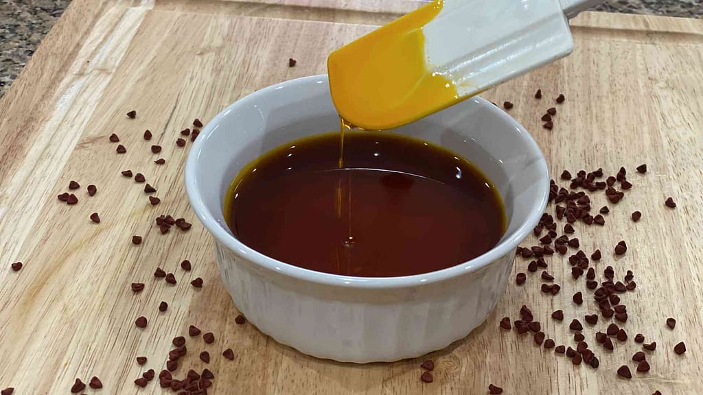 Annatto Oil in a glass bowl with annatto seeds surrounding on a wood cutting board